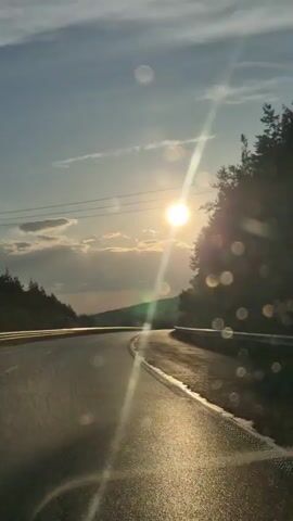This is a video of a road trip at sunset, likely in Gorno Kamartsi, Bulgaria. The sun is low in the sky, casting a warm glow on the wet asphalt road, suggesting it may have recently rained or that the road is damp from morning dew. The sky is a clear blue with scattered white clouds, and the sun's rays create a prominent lens flare effect, adding to the serene atmosphere. Trees line both sides of the road, with the right side featuring a denser forest, hinting at a rural or mountainous setting. The road itself appears to be a highway or a well-maintained rural road, curving gently to the right. There are power lines visible in the distance, indicating human infrastructure. The overall mood is peaceful and reflective, capturing the beauty of a sunset drive. There is no visible text in the media.