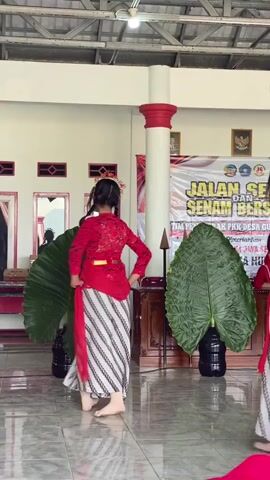 A group of young women in red tops and striped skirts are performing a dance. They are barefoot and wear traditional headdresses. The dance appears to be taking place indoors, in a room with a tiled floor and a high ceiling with visible support beams. In the background, there is a banner with Indonesian text, and two large green artificial leaves are positioned as props. There are also pillars in the room, one of which is red and white. Several framed pictures hang on the wall behind the dancers. The overall impression is of a cultural performance or event.