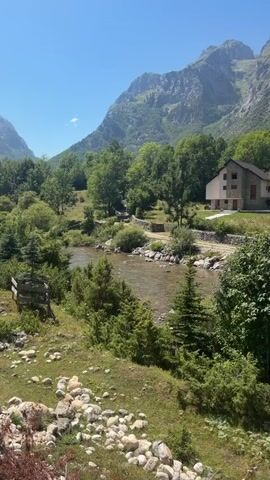 This is a vertical, handheld, mid-shot video of a tranquil mountain landscape. The video captures a clear blue sky over lush green mountains and trees. In the foreground, a narrow, clear river flows with rocks visible on its bed and along its banks. Several pine trees and shrubs are scattered throughout the scene. On the right side of the river, a dirt road winds through the trees, bordered by low stone walls. Further back, a modern house with a beige facade and a dark roof is nestled amongst the trees. The overall impression is one of serene natural beauty.