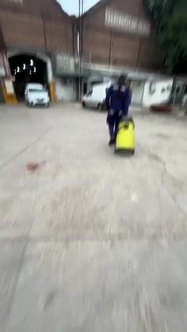 A person, seen from behind and wearing dark blue workwear, pulls a yellow Karcher pressure washer on wheels across a concrete or paved outdoor area. In the background, there is a large brick building with "QUINQUENAL" painted on its side, suggesting an industrial or old factory setting. To the left of the building, a white car is partially visible, and another white van is parked further to the right. Trees and power lines are visible above the buildings. The sky appears overcast. The scene depicts an outdoor work environment in Sáenz Peña, Argentina, likely during the day.