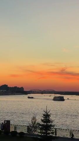 A vertical, low-angle shot shows a river at sunset. The sky is a gradient of soft blue at the top, transitioning to orange and pink clouds near the horizon. A city skyline with buildings and a bridge is visible in the distance. Several boats are on the water. In the foreground, a black metal fence runs along a walkway, behind which are a few small trees and what appears to be a trash can on the left.