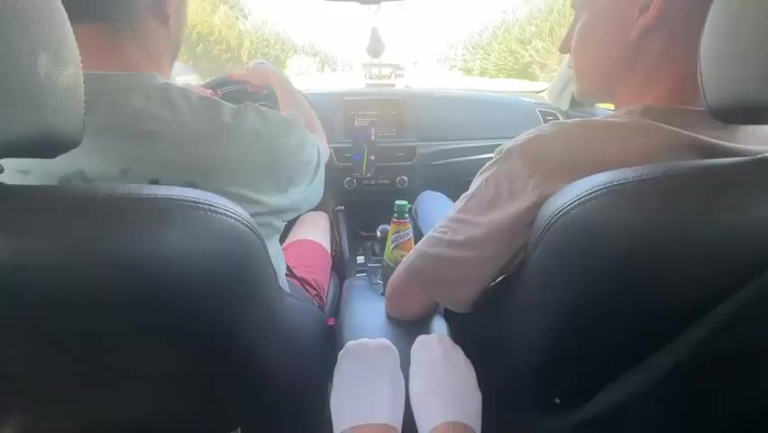 From the rear passenger seat perspective, two men are in the front seats of a car, driving down a road with trees visible through the windshield. The driver is wearing a green shirt and red shorts. The passenger is wearing a tan shirt and jeans.  A bottle of juice is visible in the center console.  Someone's feet, clad in white socks, are in the center console. The driver gestures with his hand as he talks. The dashboard and navigation screen are visible.