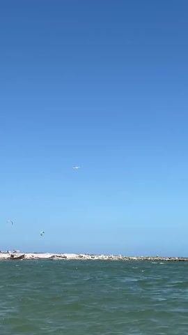 The camera pans across a scene featuring a body of water, a shoreline, and a clear blue sky. In the distance, a small airplane is visible against the blue expanse. The water, which takes up a significant portion of the frame, is green and textured with gentle waves. Along the shoreline, there are white buildings partially hidden by trees. Some people are seen swimming in the water, while others are on the beach. In the far distance, wind surfers can be seen in the sky.