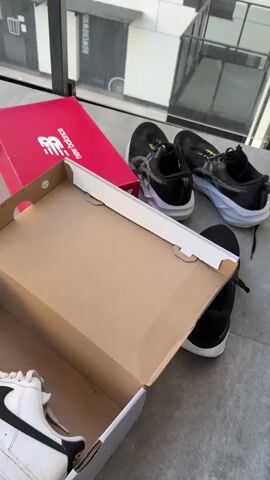 The video shows a person cleaning sneakers on a balcony. In the first frame, a red shoebox and a pair of black and white sneakers are visible on the floor, next to an open cardboard shoebox. In the second frame, a young person wearing a white t-shirt and gray shorts is holding a white sneaker, seemingly cleaning its sole with a green cloth. In the third frame, a pink bucket filled with pink liquid is on the floor, next to a pair of white sneakers. One of the white sneakers has several plastic water bottles strapped to it. In the fourth frame, the pink bucket with the pink liquid is more prominently shown. In the background of all frames, a modern building with windows and balconies is visible, along with a parking lot with cars and trees.