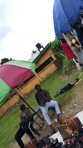 The video begins with a tilted view of an outdoor scene. A large, multi-colored umbrella, predominantly green and pink, is open and angled. Beneath the umbrella, two men are visible, sitting or crouching. One man wears a dark shirt and patterned pants, holding onto the umbrella's pole. The other man, in a dark shirt and black pants, is also near the umbrella. In the background, a building with yellow walls and a brown roof is visible, along with some trees and vegetation. The sky is cloudy. The camera then shifts to focus on a large blue umbrella, also open and angled. A woman with braided hair is partially visible in the foreground, looking down. More of the yellow-walled building and greenery are in the background. The video ends with a closer view of the blue umbrella and a glimpse of two people sitting on the ground to the right.