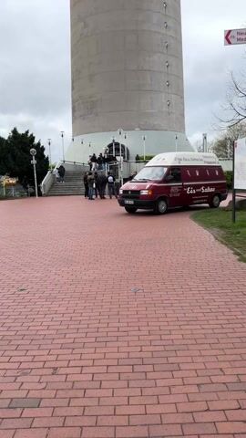 The camera pans upwards, starting from ground level, to show a very tall concrete tower that tapers slightly towards the top. At the base of the tower, a group of people are congregated near an entrance. A red van with the words "Eis und Sahne" (Ice cream and cream) written on it is parked nearby. The ground is paved with red bricks. The sky is overcast. The top of the tower has a round, glass-enclosed structure.