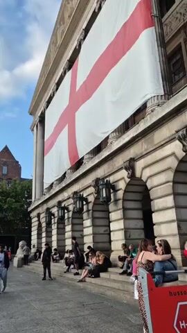 The video shows a street scene with a large English flag hanging on a building. People are sitting on the steps of the building and walking along the street. There are also market stalls and other buildings in the background. The sky is blue with some clouds.