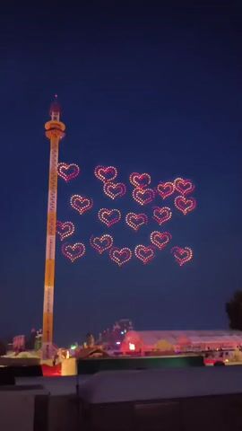 A drone show displays numerous heart-shaped formations in the night sky. The hearts are illuminated with red and white lights, forming a large cluster in the center of the frame. To the left, a tall, yellow and white tower with lights visible on its structure stands prominently. Below the drone formations and the tower, the lights of an amusement park or fairground are visible, including tents and other attractions. The sky is dark blue, indicating it is nighttime.