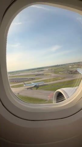 The video shows a view from an airplane window. The window frames a large airport tarmac with several airplanes visible on the ground and a long runway. Green grass surrounds the tarmac. The sky above is clear blue with some scattered white clouds. The sun appears to be shining, casting a bright light. The interior of the airplane is visible around the window, with the curved white wall of the cabin and a portion of an engine. The perspective suggests the plane is either taxiing on the ground or has just taken off or landed.