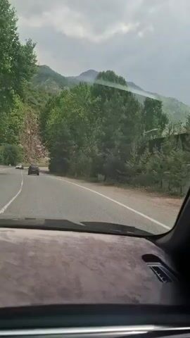 The video shows a car driving on a two-lane road through a mountainous, wooded area. Two cars are ahead of the camera car. The road curves around a rock face and through trees. The sky is overcast. The inside of the camera car has a gray dashboard. Music is playing in the background.