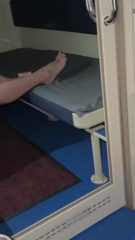 The video shows the inside of a sleeping cabin on a train. A woman is lying on the bottom bunk. She is lying on her side and looking at her phone, which she is holding up in her right hand. Her bare feet are resting on the edge of the bunk. There is a pink bag next to her. The cabin is small and has a blue floor with a red rug. There is a window with a reflection visible, showing a water bottle and some snacks. The door to the cabin is partially open.