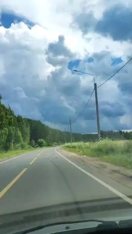The video shows a point-of-view shot from a moving vehicle on a paved road with a yellow line on the left and a white line on the right. The road curves to the left and is bordered by lush green trees and vegetation. The sky is dramatic with large, dark clouds and patches of blue sky, suggesting an overcast or impending storm. Several utility poles with streetlights and overhead wires are visible along the side of the road. The overall scene depicts a drive through a rural or semi-rural area under a dynamic sky.