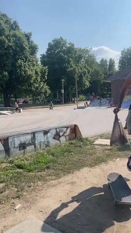 The video shows a skatepark on a sunny day with several people present. In the background, trees and buildings are visible. The foreground shows a dirt path with grass and some discarded items, including skateboards and scooters.

The skatepark features ramps, ledges, and other obstacles. Some people are seen riding bikes and scooters. One person is wearing a safety vest and appears to be working on the park. Another person is seen near a fence with a broom.

The video captures the activity and atmosphere of a public skatepark, highlighting the recreational use of the space and the various elements that constitute it.