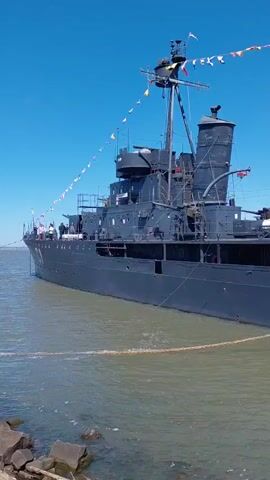 A medium-sized, dark gray naval vessel is docked in a body of water under a clear blue sky. The ship is adorned with a string of colorful nautical flags. The camera pans across the side of the ship, showing its deck, a mast with more flags, a smokestack, and what appears to be a cannon mounted on the deck. A rope is visible in the foreground, stretching from the shore to the ship. The water is calm and appears slightly murky. The video is taken from a low angle, near the water's edge.