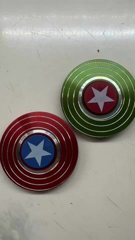 Two fidget spinners sit on a white surface. The spinner on the left is red with a blue center and a white star. The spinner on the right is green with a red center and a white star. Both spinners are not in motion. The surface has several scratches.