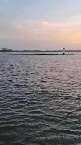 The video shows a wide body of water with a lighthouse at the end of a long pier in the distance. The sky is hazy with pastel colors, suggesting either dawn or dusk. There are a few indistinct figures visible on the pier, some near the lighthouse and others spread out along its length. Buildings can be seen on the far left horizon. The water is choppy with small waves. The pier extends approximately 400 meters into the water.