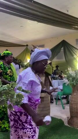 The video shows a woman dancing in what appears to be a hall or event space. She wears glasses, a light gray headwrap, a white and lilac lace top, and a purple and white patterned skirt. She holds a small white object, possibly a fan or tissue, in her right hand. Around her are other people dressed in similar festive attire, suggesting they are at a celebration, possibly a wedding or similar event. Green and white fabric is draped from the ceiling and behind the people at tables set in the background, possibly to decorate the space. There are also plants and woven baskets adding to the decor. The lighting is bright, giving the scene a cheerful atmosphere.