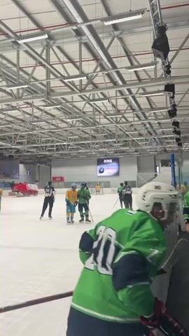 This media file depicts a hockey game taking place in an indoor ice rink. The scene is brightly lit, indicating daytime or the use of artificial lighting within the arena. Several individuals are visible on the ice, including players in green and yellow uniforms, and at least one referee in black and white stripes. They are all wearing hockey gear, including skates and helmets, and some are holding hockey sticks. The ice surface shows signs of use, with visible skate marks. In the background, there is a large scoreboard displaying a timer that reads "02:52." The setting is a modern indoor sports facility with a high, metallic ceiling structure and industrial-style lighting. There are also bleachers or seating areas visible along the side of the rink. The overall atmosphere appears to be that of a competitive sporting event. Based on the provided location context, the scene is in Qalansawe, Israel.
