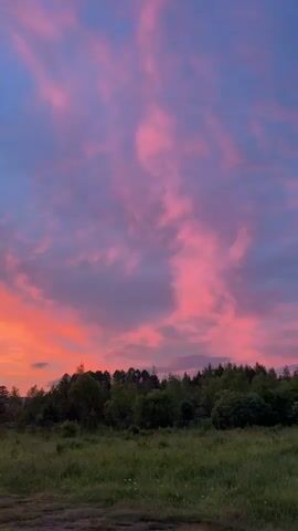 The video shows a sunset over a landscape of rolling hills and trees. The sky is filled with vibrant colors of pink, orange, and purple, with clouds streaking across the sky. The foreground is a grassy field, with a dirt path winding through it. The overall impression is one of natural beauty and tranquility.
