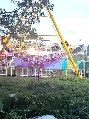 The video captures an outdoor amusement park scene at dusk in Matesih, Indonesia. The primary subject is a pirate ship amusement ride, brightly illuminated with numerous colorful lights, predominantly purple and white/yellow. The ride is actively swinging back and forth. In the foreground, there are trees with dark foliage and an area of green grass and smaller plants, separated from the ride by a low white fence. As the video progresses, the sky visibly darkens from a lighter blue to a deeper twilight hue, making the ride's lights stand out more prominently. No specific people are clearly visible, but their presence is implied by the active ride. There is no discernible text.