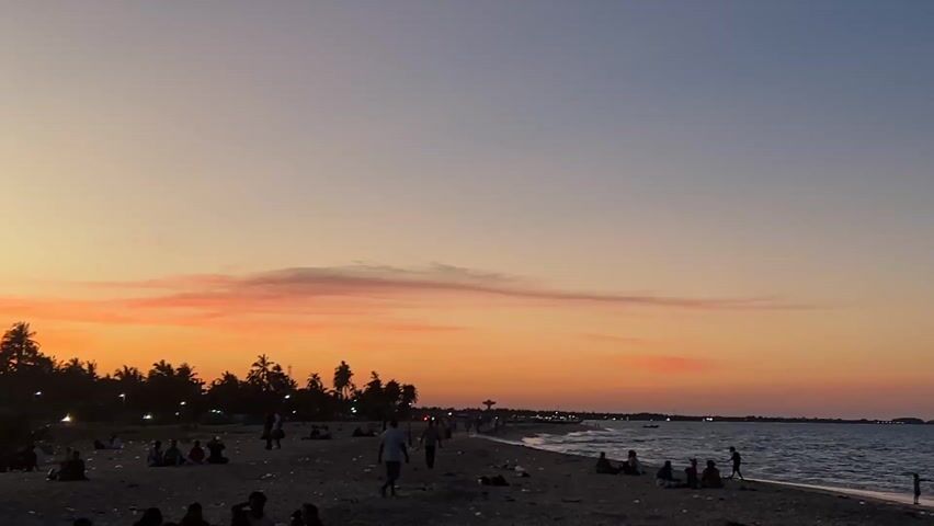 The video captures a serene sunset on a beach in Kalmunai, Sri Lanka. The sky presents a striking gradient, transitioning from vibrant orange and red hues near the horizon to a softer pale blue-grey at the top, with wispy clouds illuminated in warm pinks.

In the foreground, a dark sandy beach is dotted with numerous people enjoying the evening. Most individuals are seated in small groups, relaxing and observing the sunset, while a few figures are seen standing or walking casually along the shore. Gentle waves are visible breaking on the right side of the frame.

A dense line of silhouetted palm trees stretches across the middle ground, creating a distinct horizon against the colorful sky. Behind these trees, faint lights from buildings, streetlights, and vehicles with headlights on indicate the presence of a coastal road or settlement. On the far left, a prominent street light and a vendor's stall displaying red and white balloons are visible, adding to the local scene.

The activity primarily involves people relaxing and enjoying the tranquil evening on the beach. The time of day is clearly sunset, marked by the dramatic sky colors. The setting suggests a peaceful coastal environment. No visible text is present in the video.