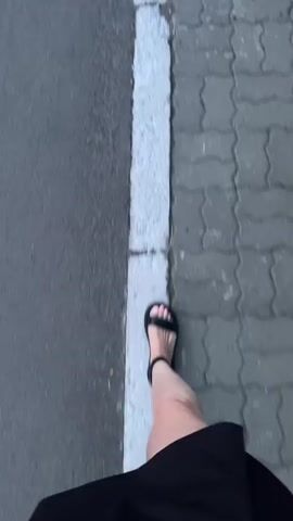The video shows a first-person perspective of someone walking along a paved surface with a white line separating it from a darker asphalt area. The person is wearing black sandals and a black skirt or dress. The camera is angled downwards, focusing on the feet and the ground. The paving stones are a light grey interlocking brick pattern. The asphalt is a dark grey, wet or damp in appearance. The white line is thick and appears to be painted. The movement suggests walking at a normal pace.