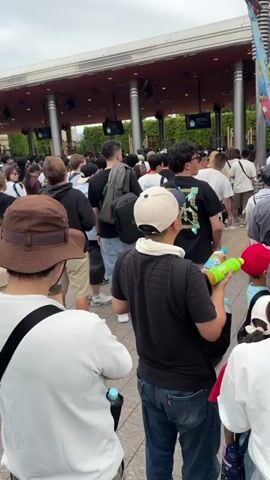 The video captures a large crowd of people gathered at the entrance of what appears to be an amusement park or similar attraction in Osaka, Japan. The sky is overcast, indicating daytime. People of various ages, including adults and children, are visible, many carrying backpacks or bags, suggesting they are visitors. Some individuals are seen holding water bottles, and one child is in a stroller.

The entrance features a large, modern archway structure, with a partially visible sign that reads "WELCOME." Utility poles and streetlights are also present, integrated into the architecture. The crowd is densely packed, suggesting a popular destination or a busy time. People generally appear to be waiting or queuing, looking forward or observing their surroundings. One person's white t-shirt has partially readable text on the back: "...CULTIVATES LOVE DOES NOT... LOVE DOES NOT CULTIVATES..." The overall atmosphere suggests anticipation and readiness to enter the venue.