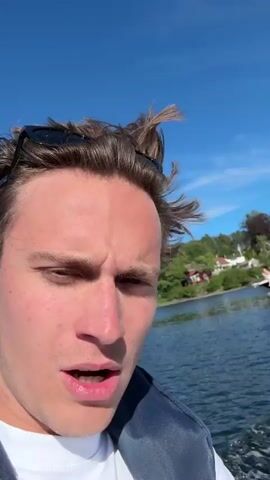 Two young men are on a motorboat on a sunny day. The man in the foreground is facing the camera and speaking, with his hair blowing in the wind. He is wearing a white t-shirt and a blue life vest. The man in the background is steering the boat and wearing a blue baseball cap, a white t-shirt, and a black life vest. They are on a body of water, with trees and houses visible on the shoreline in the distance. The boat is creating a wake behind it.