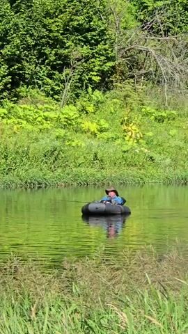 A person wearing a hat and a blue shirt sits in an inflatable black raft on a body of water. The person is holding a fishing rod and appears to be fishing. The water is green and reflects the surrounding trees and foliage, which are also green. The scene is lit by sunlight.