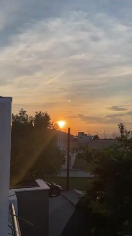 The video captures a sunset viewed from a rooftop. The sun is a bright orange orb low in the sky, casting golden rays through the clouds. The sky is a mix of pale blue and orange hues, with streaks of white clouds. In the foreground, a white structure, possibly part of a building, is visible on the left. Dark green foliage from trees frames the right side of the image. In the middle ground, there are houses and buildings, and a dark-colored car is parked near the trees. A telephone pole stands vertically in the scene. The overall environment appears to be a residential area.