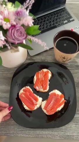 A close-up shot shows a black plate with three open-faced sandwiches. Each sandwich consists of a slice of toasted bread topped with cream cheese and slices of smoked salmon. To the left of the plate, a woman's hand with red nail polish is visible, and to the right, a mug filled with dark liquid, likely coffee, is present. In the background, a laptop and a vase containing pink flowers and greenery are arranged on a wooden surface. The lighting is soft, suggesting an indoor setting, possibly a desk or table.