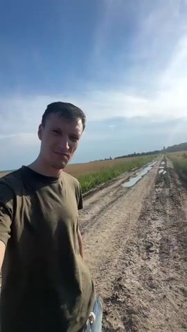A man in a green t-shirt is standing on a dirt road next to a field of wheat. The road has puddles of water on it and tire tracks. The man is smiling and holding up two fingers in a peace sign. He is in the foreground of the video. In the background, there is a field of wheat and a blue sky with white clouds. The video then pans to show a destroyed car in the field of wheat. The car is rusted and burned out, and it is overgrown with weeds.