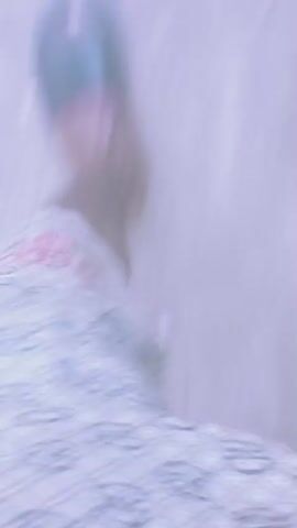 The video shows a first-person perspective of someone walking on a textured surface, possibly dirt or gravel. The footage is blurry and in motion, making it difficult to discern clear details. Several blurry figures can be seen in the background, some appearing to be wearing dark clothing. The person is wearing footwear, possibly sandals, with a patterned fabric visible. The lighting is soft and diffused, with a pale, muted color palette dominating the scene. There are occasional specks of lighter material scattered on the ground. The overall impression is of movement and a slightly overcast environment.