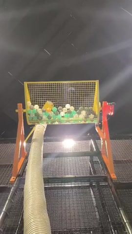 This is a low-angle shot looking up at a ball pit machine. A large, flexible tube extends from the bottom of the frame upwards towards a yellow mesh bin filled with green, white, and black balls. The bin is mounted on an orange metal frame. Light shines from behind the bin, illuminating the balls and creating lens flares. As the video progresses, balls are blown out of the bin through the tube and into the air, creating a flurry of motion. Some balls appear to be coming directly from the tube, while others are being ejected from the bin itself. The background is dark and indistinct, suggesting an indoor play area.