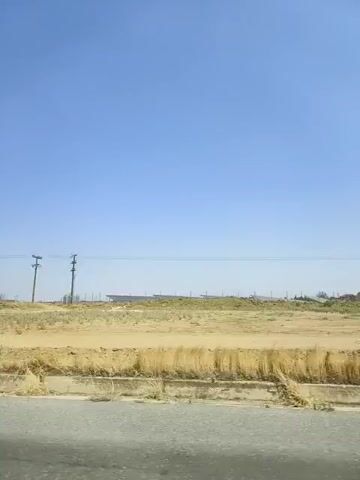 The video shows a barren landscape with dry grass and dirt under a clear blue sky. There are power lines and utility poles visible. The camera appears to be moving, possibly from a vehicle, as indicated by the slight motion blur and the perspective of the road or ground in the foreground. The scene is rural and appears to be a dry, possibly arid, environment. There are no people or animals visible in the video. The overall impression is one of a vast, empty landscape.