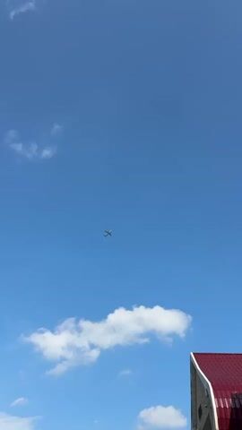 A video shows a bright blue sky with a few scattered white clouds. A small airplane is visible in the center of the frame, flying against the blue sky. In the bottom right corner of the frame, the corner of a building with a red roof is visible. The video is short and appears to be a single, static shot. There are no people or discernable interactions visible in the video. The environment is an outdoor setting during the daytime.