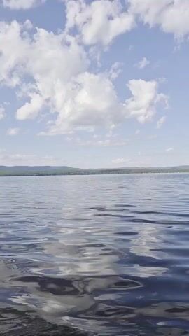 The video shows a vast body of water, likely a lake, under a bright blue sky filled with scattered white clouds. The water's surface is gently rippled, reflecting the sky and the distant, tree-covered shore. The horizon is visible in the distance, where the water meets the land and the sky. The scene is tranquil and evokes a sense of natural serenity.