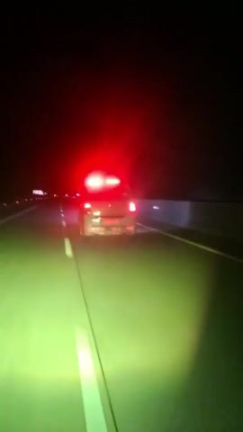 The video captures a view from a vehicle driving on a highway at night. The road is illuminated by the vehicle's headlights, casting a green glow. Ahead, another vehicle is visible, its red taillights and white headlights are illuminated. The vehicle appears to be a white van. The road has white lane markings. The sky is dark, and the surrounding environment is not clearly visible. The video captures the movement of the vehicles on the highway.