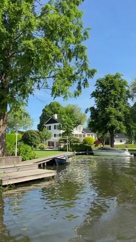 The video captures a scenic view from a boat on a canal. The camera pans across the lush landscape, revealing tall green trees, reeds along the water's edge, and glimpses of houses with docks. The water is calm, reflecting the clear blue sky. The focus is on the natural beauty of the surroundings, with the sound of the boat audible in the background.