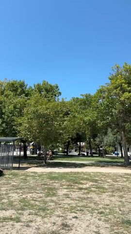 The video shows a clear blue sky with a few trees and some buildings in the background. There is a sandy area with some beach umbrellas and chairs, suggesting a beach or park setting. A person is seen walking in the distance. The video pans to show more of the beach area with people relaxing on the sand and in the water. There are also mountains visible in the background. The overall scene depicts a sunny day at the beach.