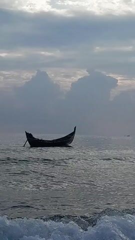 A person rows a small wooden boat in a body of water. The boat is dark and appears to be a traditional design, with raised prows at the bow and stern. The person is silhouetted against the cloudy sky, and the water is choppy with small waves. Other boats are visible in the distance. The sky is overcast with thick, grey clouds.