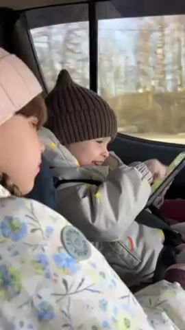 Two young children, likely siblings, are sitting in the back seat of a moving car. The child on the right, wearing a brown pointed hat and a light-colored jacket with a rainbow design, is engaged with a tablet, pointing and smiling. The other child, wearing a pink hat and a floral jacket, is looking towards the tablet with a neutral expression. Both children are buckled into their seats. The car window shows a blurred landscape of trees and buildings passing by.