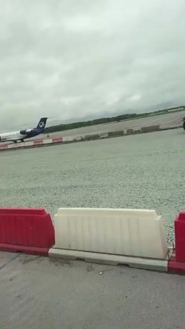 The video shows an airplane on a tarmac at an airport under a cloudy sky. The airplane is white with blue and dark blue markings and the logo "OK" on its tail. Two people are visible near the front of the airplane, appearing to be airport personnel. In the foreground, there are red and white plastic barriers. A white vehicle is partially visible on the left side of the frame. The airplane is positioned on a runway or taxiway, with checkered red and white markings visible on the ground. The area around the runway appears to be covered in gravel or small stones. The camera is positioned low, likely from the perspective of someone sitting in a vehicle.