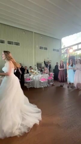In a large room with light-colored walls and a white ceiling, a woman in a white wedding dress with a full skirt and train is positioned on the left side of the frame, facing away from the camera and holding a bouquet of flowers. Her hair is blonde and is tied back. She appears to be in motion, possibly turning or preparing to throw the bouquet. Behind her, a table is set with white tablecloths, pink floral arrangements, and chairs. Guests are seated at the table, and some are standing. In the background, several women in formal dresses are standing on the right side of the frame, facing the woman in the wedding dress. One woman in a dark red dress, one in a light blue dress, and another in a light pink dress are clearly visible, along with others partially obscured. A man in a dark suit is standing near the table and appears to be speaking or gesturing. Further back, a DJ booth with lighting equipment is visible. The floor is made of polished wood. The overall atmosphere suggests a wedding reception or a similar celebratory event.