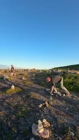 This video shows two people in an outdoor rocky landscape with sparse vegetation. In the foreground, one person wearing a red cap and dark clothing is bending down and appears to be interacting with rocks. Another person, wearing a light-colored outfit and hat, stands a short distance away, looking towards the person bending down. The landscape is dotted with numerous small rock cairns, suggesting a possible hiking trail or a place of reflection. In the background, a car is visible on a road, and a vast, open, and hilly terrain stretches out under a clear blue sky. The lighting suggests it is either sunrise or sunset, with a warm glow on the horizon in some frames. The overall mood appears calm and serene, with the vastness of the landscape and the simple act of arranging rocks creating a sense of peacefulness. There is no visible text in the video.