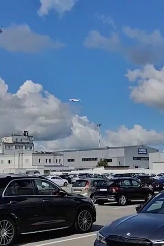 A distant airplane flies across a cloudy blue sky over an airport tarmac. In the foreground, several black cars are parked in a lot. A tall tower with lights and antennas stands in the middle of the frame.