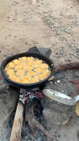 The video shows a black cast iron pan filled with food items being fried in oil over an open fire. The food items are golden brown and appear to be small, round fritters or dumplings, with several pieces submerged in the bubbling oil. The pan is resting on stones, which are part of a makeshift cooking setup on sandy, gravelly ground. Embers and logs are visible beneath the pan, indicating the heat source. A metal slotted spoon rests on a stone beside the pan. The overall scene depicts an outdoor cooking scenario, likely in a rustic or camping environment.