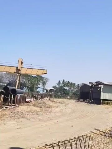 The video pans across an outdoor scene under a clear blue sky. In the foreground, a chain-link fence runs horizontally. To the left of the fence is a dusty, unpaved area with some scattered debris and what appears to be construction materials or equipment. A large yellow crane-like structure is partially visible in the far left of the frame. Behind the dusty area, there are several simple buildings with corrugated metal siding. To the right of the dusty area, a paved walkway runs alongside a white building with a dark roof. Several air conditioning units are mounted below the windows of the white building. A date palm tree and other green foliage are planted between the fence and the walkway, near the white building. The overall impression is an industrial or semi-developed outdoor area, possibly a construction site or a rural facility.