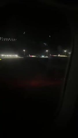 The video shows a view from an airplane window at night. The runway lights and other airport lights are visible in the distance, along with several parked airplanes. The plane appears to be moving, as indicated by the slight blur of the lights. The interior of the airplane is dark, with only the reflection of the lights on the window visible. The video is shaky, suggesting it was filmed by a person holding a camera or phone.