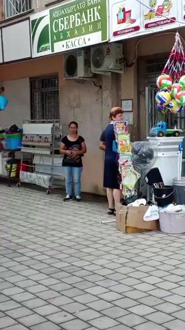 A pavement is visible in the foreground with grey and white square tiles. Two women are standing in front of a shop. The woman on the left is wearing a black t-shirt and light blue jeans. The woman on the right is wearing a dark blue dress. A shelf with many magazines is in front of the woman on the right. Various goods are displayed outside the shop including toys such as a blue dolphin balloon, red and green balls in a net bag, and a toy truck. There is also a rack with rolls of patterned paper. A sign above the shop reads "СБЕРБАНК" which is a Russian bank. An air conditioning unit is mounted on the wall above a window.